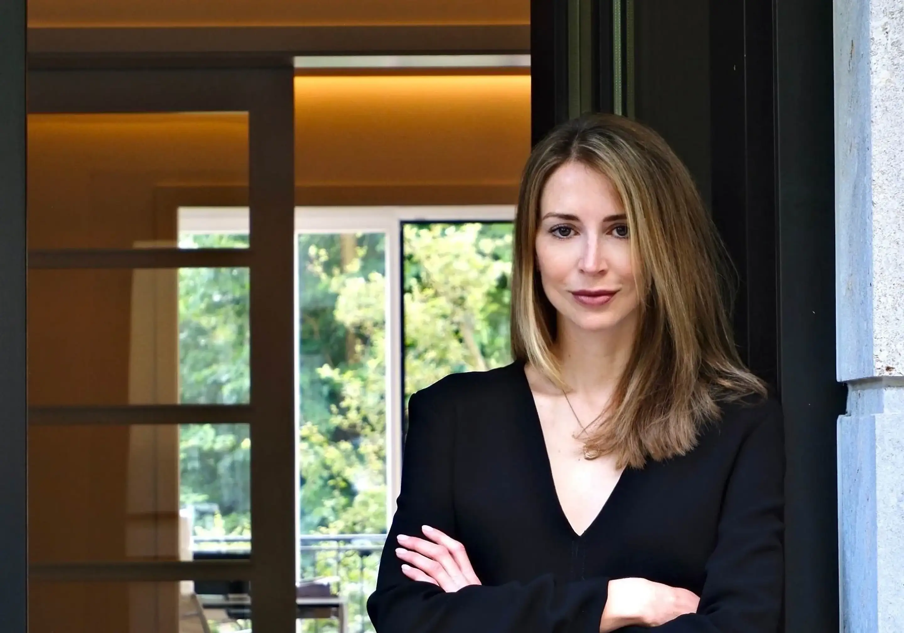 Dr. med. Lucija Grittern mit verschränkten Armen im schwarzen Oberteil vor einem Fenster mit Blick auf grüne Bäume.