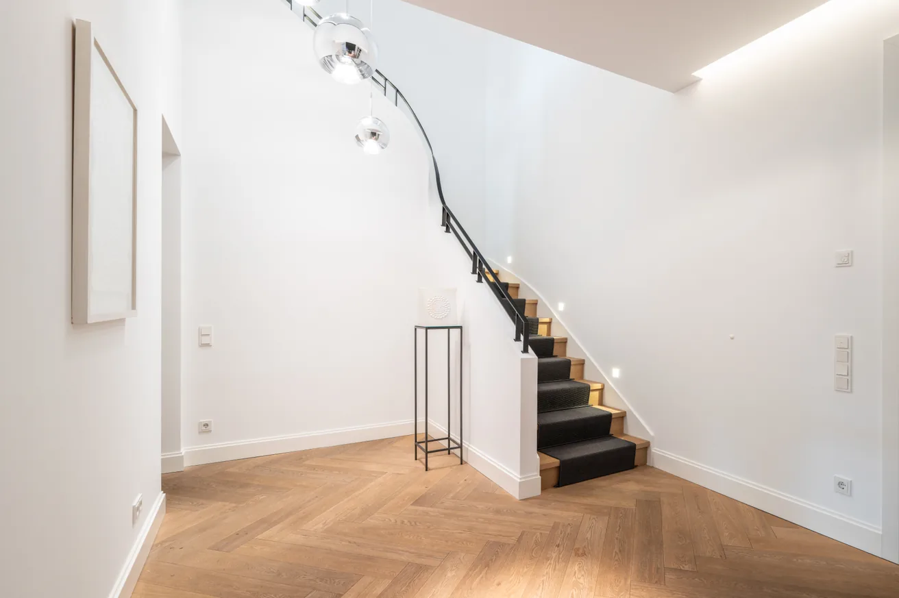 Heller Innenraum mit Holztreppe, schwarzem Treppenteppich, weißen Wänden und hölzernem Fischgrätparkettboden.
