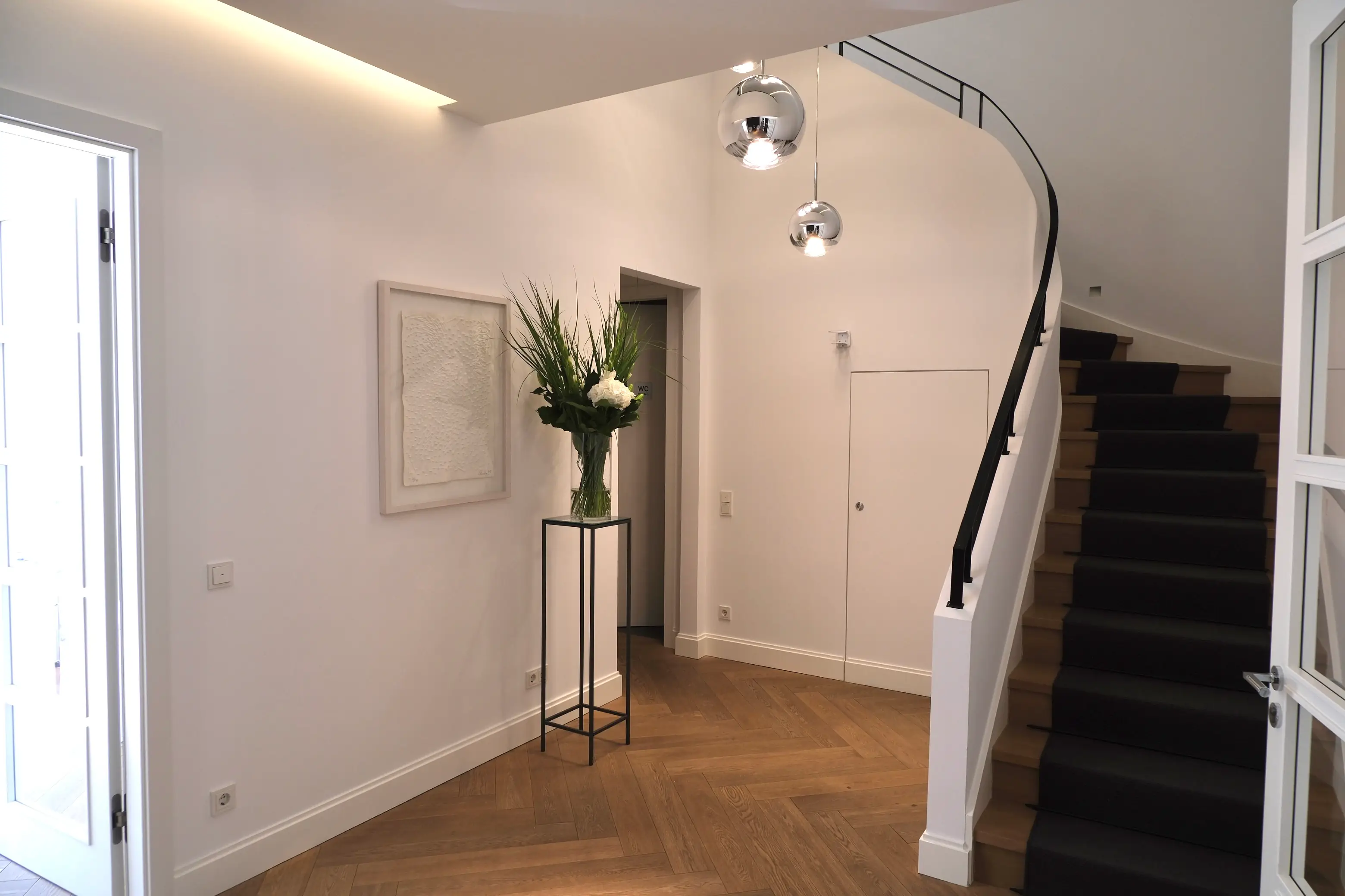 Heller Praxisflur mit Holzparkett, Treppe mit schwarzem Läufer, großem Blumenarrangement und Kugelleuchten.